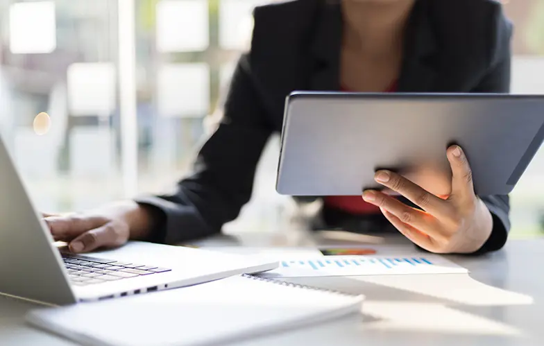 Women at desk typing on laptop while holding a tablet